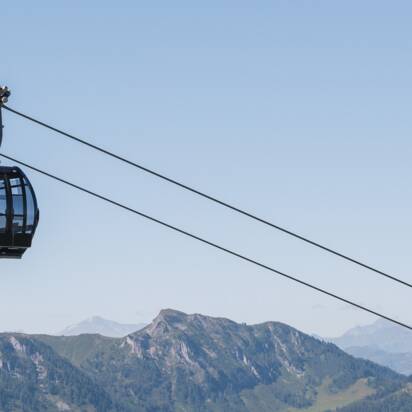 Zwei schwarze Panoramagondeln schweben über grünen Bergen im Sommer, im Hintergrund schneebedeckte Alpengipfel. | © Snow Space Salzburg