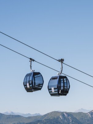 Zwei schwarze Panoramagondeln schweben über grünen Bergen im Sommer, im Hintergrund schneebedeckte Alpengipfel. | © Snow Space Salzburg