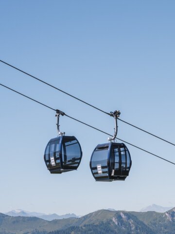 Two black panoramic gondolas float above green summer mountains, with snowy Alpine peaks in the far background. | © Snow Space Salzburg
