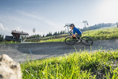 Ein Junge mit Helm fährt auf einem Woom-Bike einen Kurventrail entlang, dahinter ein Almhaus, Wiese und Gondelbahn. | © Snow Space Salzburg - Chris Eder