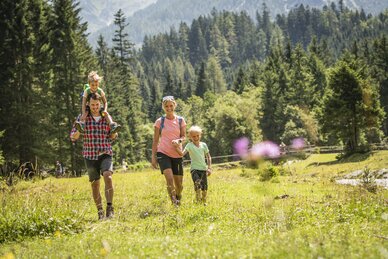 Fröhliche Familie wandert über blühende Almwiese am Waldrand im Sonnenschein bei Sommerurlaub in den Alpen.
