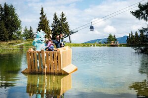 Family with mascot on wooden raft in mountain lake, gondola in background, trees and mountains visible in sunny weather | © Snow Space Salzburg