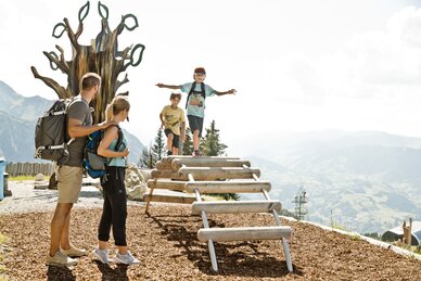 Zwei Kinder auf Klettergerüst aus Holz, Eltern beobachten sie, im Hintergrund Berge, Bäume und eine große Holzskulptur | © sanktjohann.com, Mirja Geh