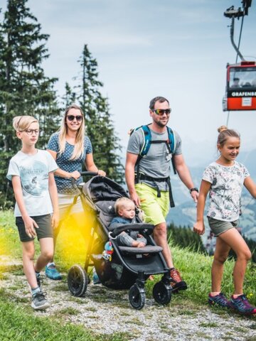 Familie mit Kinderwagen wandert am Grafenberg entlang, im Hintergrund fährt eine rote Gondel der Seilbahn vorbei | © Snow Space Salzburg