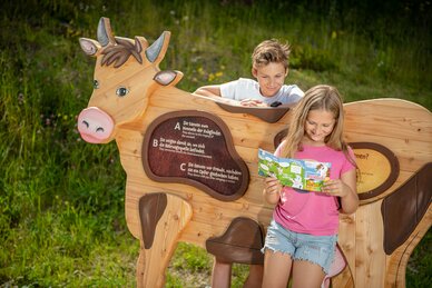Mädchen liest KUHbidu-Heft, Junge lehnt an hölzerner Kuh mit Quizfrage, grüne Wiese und Blumen im Hintergrund | © Snow Space Salzburg, Christoph Huber