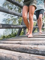 Zwei Personen gehen barfuß über eine Holzbrücke, Nahaufnahme der Beine, im Hintergrund Wiese, Wald und Wanderer | © Snow Space Salzburg