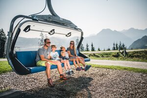 Familie mit drei Kindern sitzt auf Sesselliftbank am Boden, sonniges Wetter, Berge und Nadelbäume im Hintergrund sichtbar | © Snow Space Salzburg