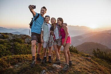 Family of four taking a selfie while hiking, sunset behind silhouettes of mountains in the scenic background