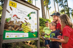 Familie mit drei Kindern steht vor Waldtafel mit Zapfenrätsel, alle halten Quizhefte, lachend und aufmerksam beim Mitraten
