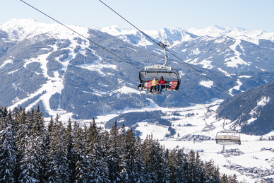 Ein Sessellift mit vier Personen schwebt über Piste mit Blick auf verschneite Berge