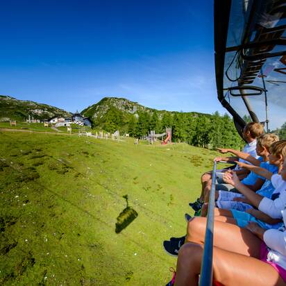 Family on summer chairlift points to mountain hut and adventure playground near Gamskogel in Zauchensee Alps.