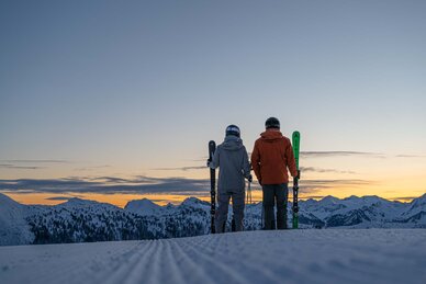 Two skiers hold their skis in their hands and look out at the sunrise