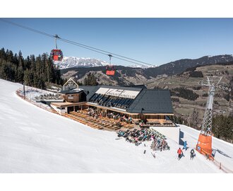 Mountain lodge with sun terrace on a ski slope, red gondola cabins above and snow-covered peaks behind