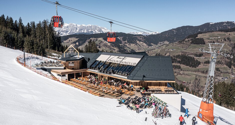 Mountain lodge with sun terrace on a ski slope, red gondola cabins above and snow-covered peaks behind