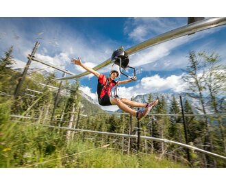 Ein junger Mann saust mit dem Flying Coaster durch den Wald auf einer Schienenbahn mit Blick auf Berge und blauen Himmel | © Christoph Huber