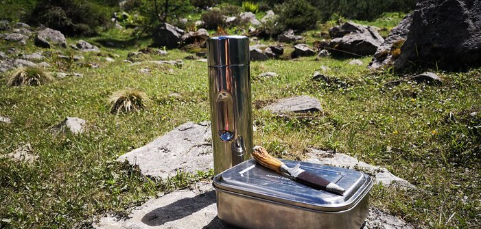 Metal lunchbox, knife and thermos bottle placed on rock in alpine meadow