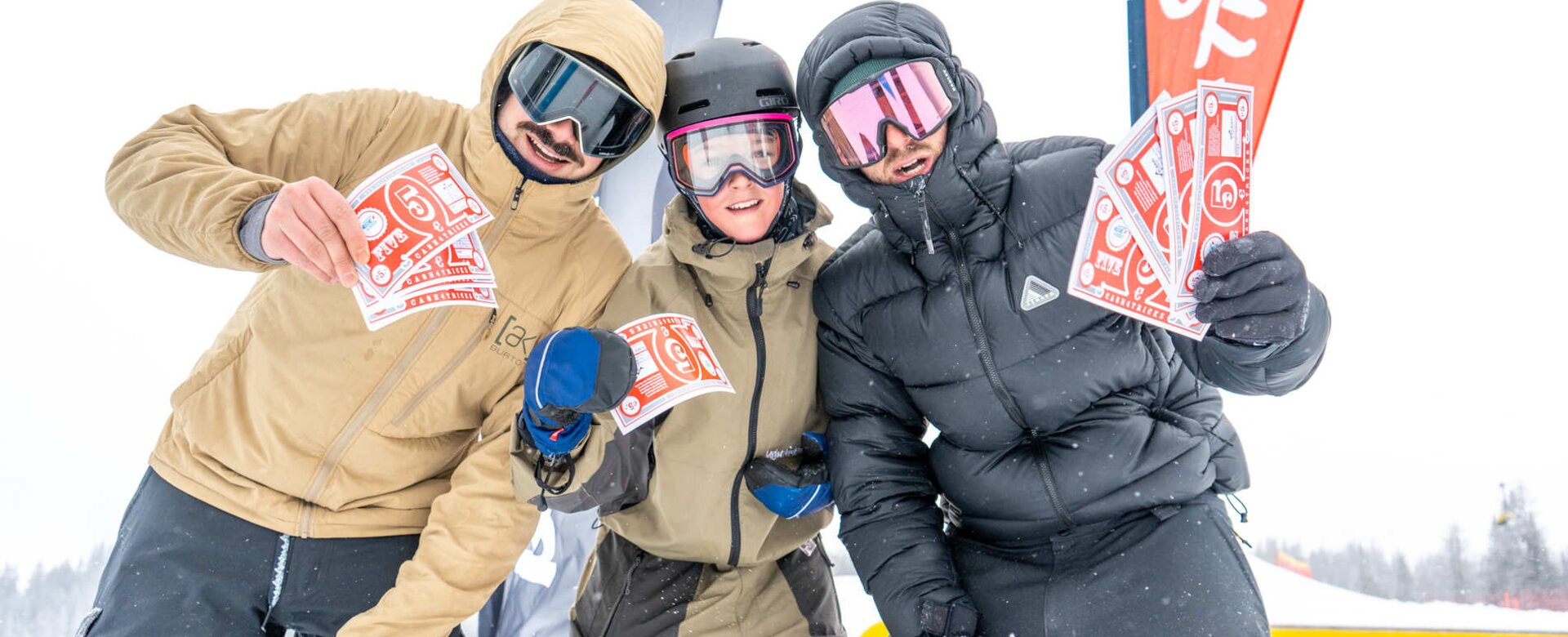 Drei Snowboarder halten bei Schneefall rote Gewinnscheine vor Fahnen im Superpark Planai in die Kamera | © Jordan Sitzwohl