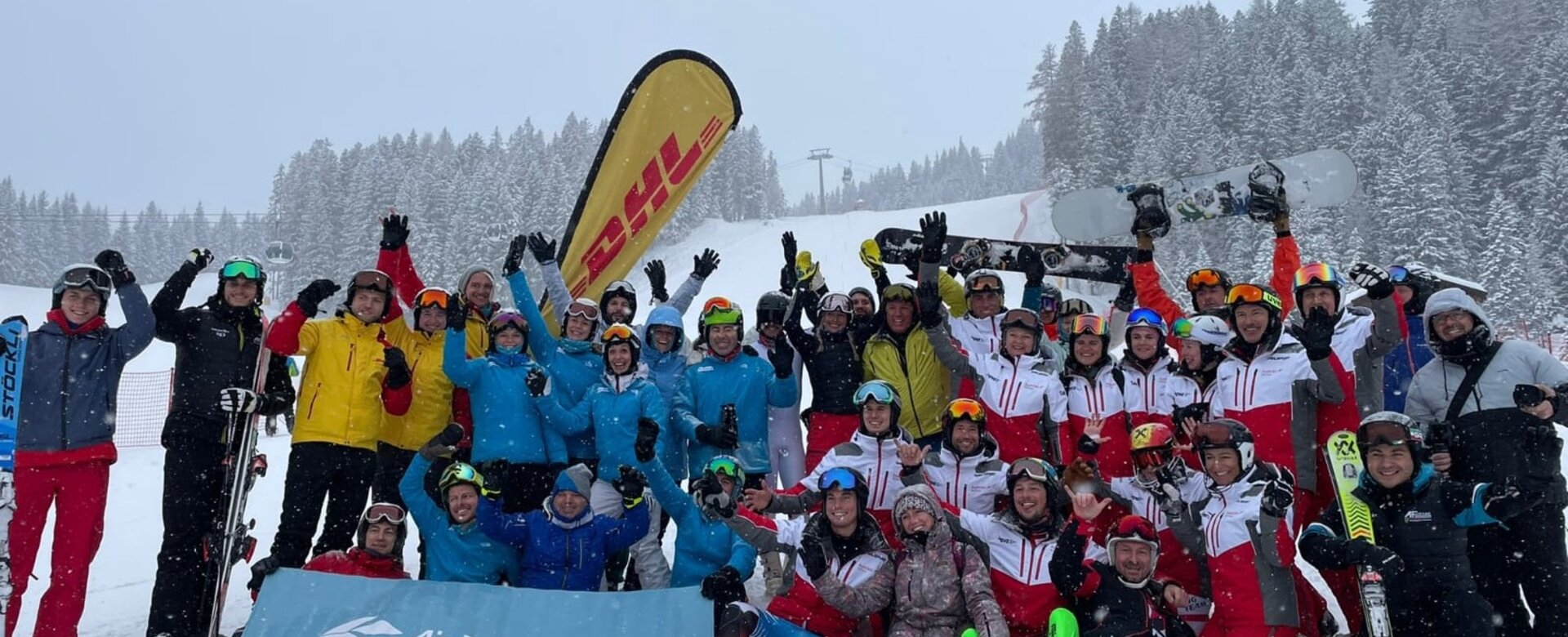 Große Skigruppe in bunter Kleidung posiert lachend im Schnee vor bewaldeter Piste