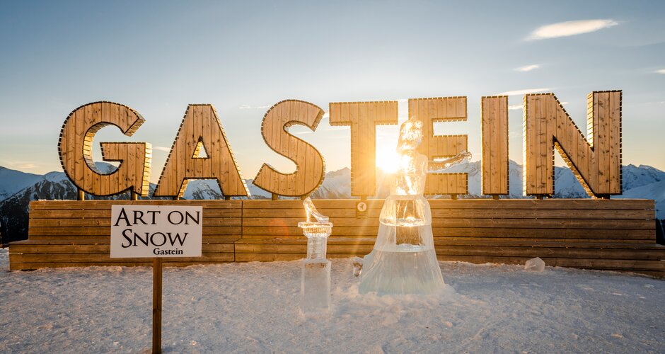Zwei Eisskulpturen stehen bei Sonnenuntergang vor großem Gastein-Schriftzug im Schnee platziert. | © TVB Dorfgastein, Max Steinbauer