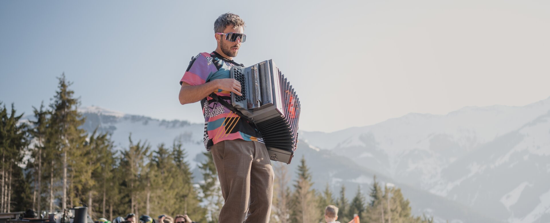 Man with accordion plays on table among cheerful guests on sunny terrace with mountain view