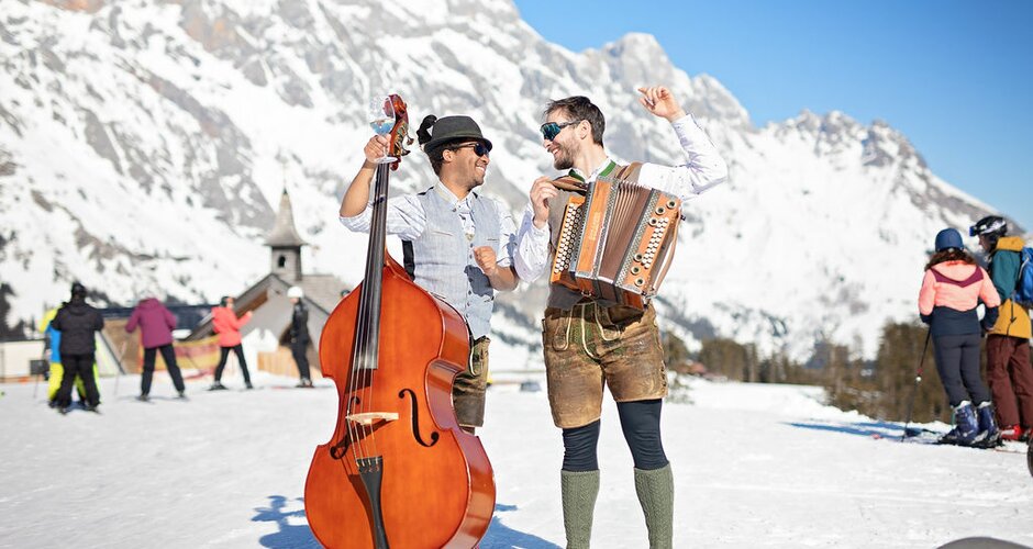 A man with a double bass and a man with a harmonica stand in the snow in front of a breathtaking panorama with the Hochkönig massif in the background. | © Hochkönig Tourismus GmbH