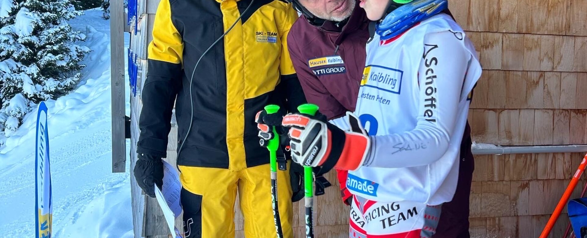 A child in ski racing gear stands in a starting gate while an adult looks at him and the other in the background smiles into his headset | © Hauser Kaibling