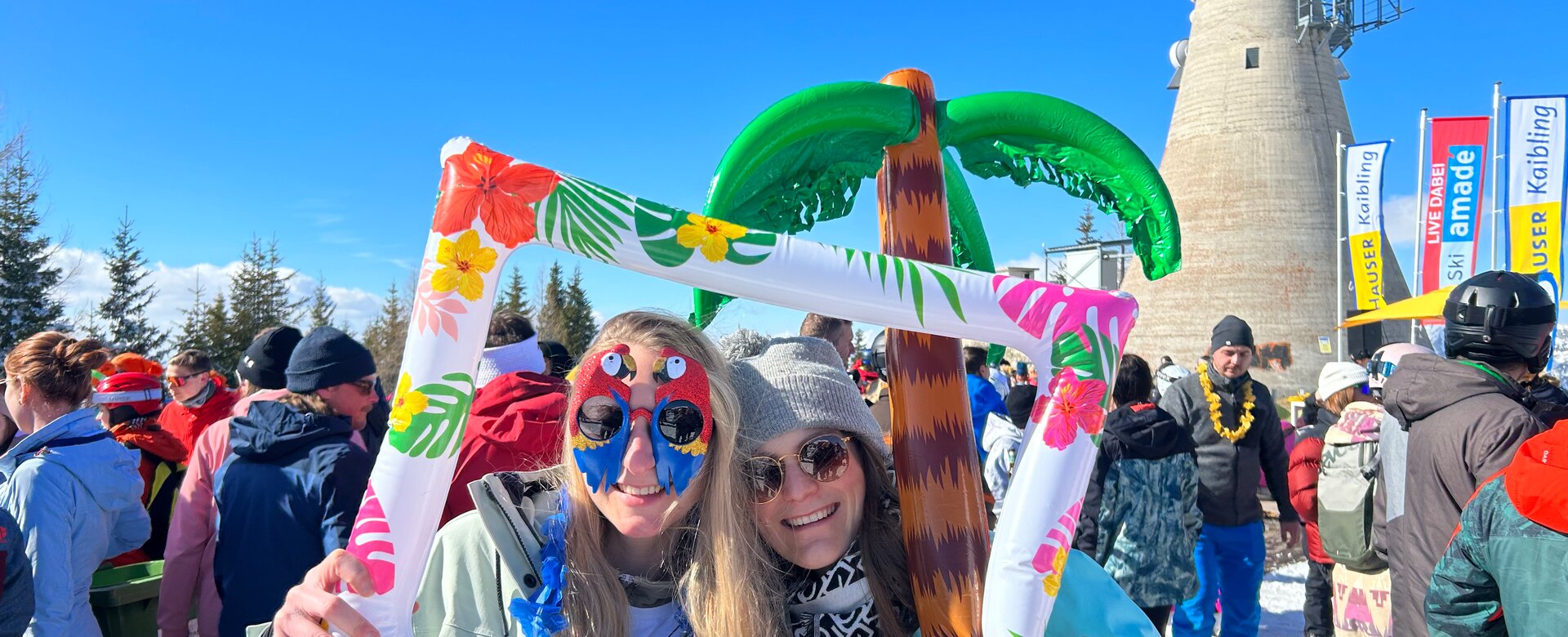 Zwei junge Damen mit winterlichem Sommeroutfit lachen in die Kamera mit einer Palme im Hintergrund. | © Hauser Kaibling