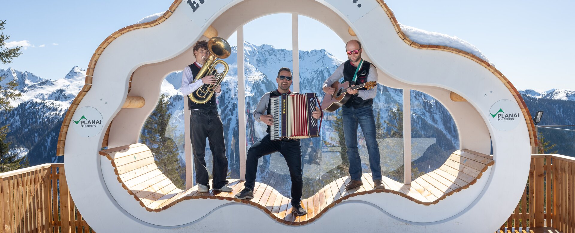 Three musicians with tuba, accordion and guitar perform on wooden platform in snowy mountain scene | © Harald Steiner