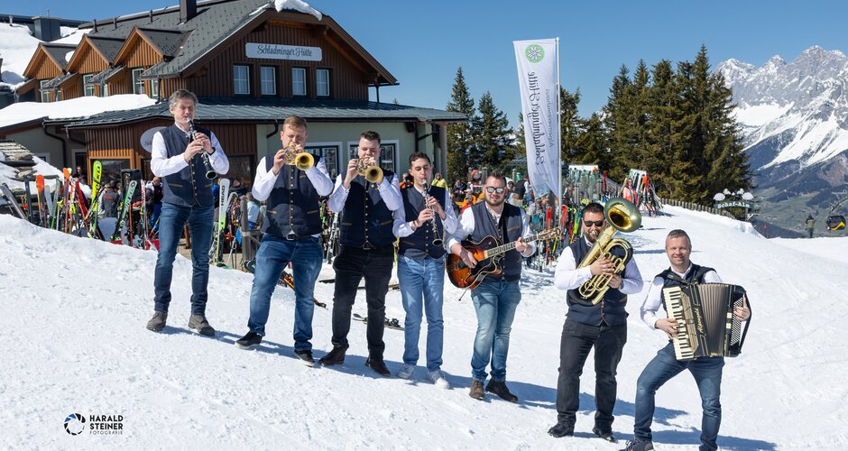 Seven musicians with instruments stand in the snow by Schladminger Hütte in bright sunshine | © Harald Steiner
