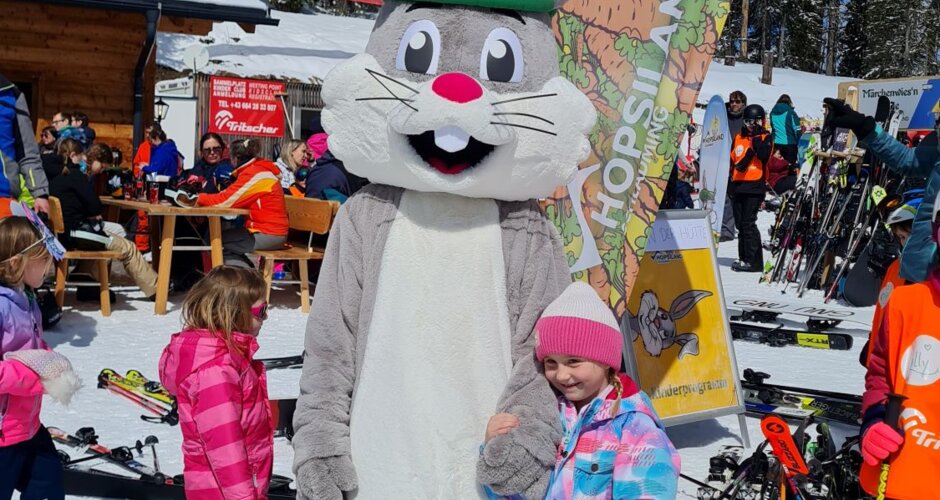 Ein Mädchen steht neben Hopsi und hält seine Hand während rund herum das Familienfest stattfindet | © Planai