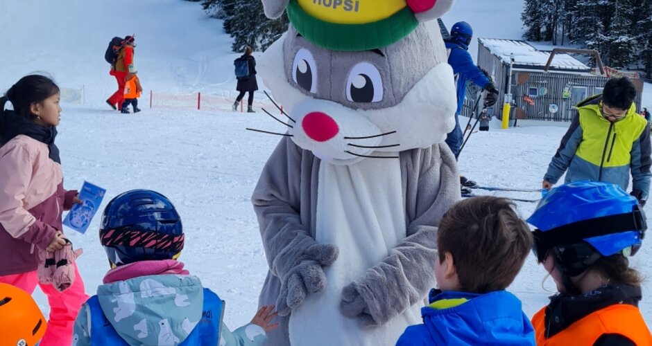 Das Maskottchen Hopsi steht inmitten von Kindern in Skiausrüstung und blickt zu ihnen hinunter | © Planai