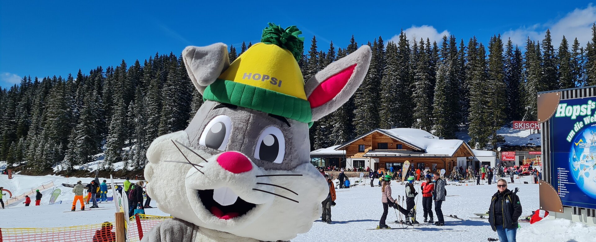 Hopsi das Hasen-Maskottchen lächelt in die Kamera und hinter ihm findet das Familienfest statt | © Planai