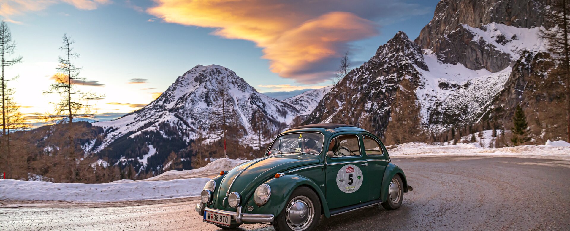 Ein grüner VW-Käfer fährt bei Sonnenuntergang auf verschneiter Passstraße vor Alpenpanorama | © Ondrey Kroutil