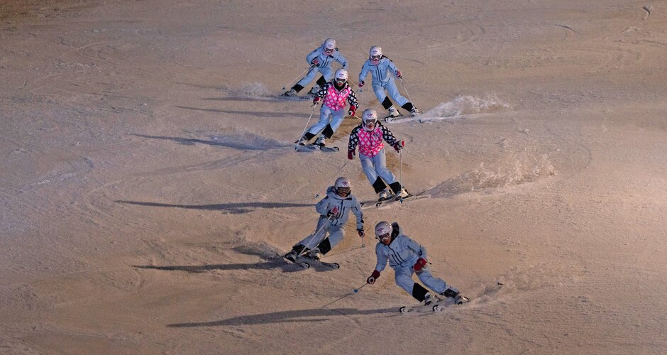 Seven skiers in formation skiing downhill on a slope lit by artificial evening light | © Michael Simonlehner