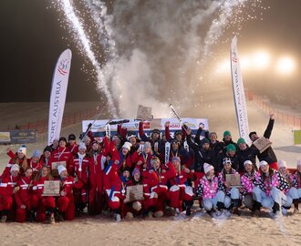 Große Siegergruppe posiert vor Feuerwerk und Bannern auf nächtlicher Skipiste | © Michael Simonlehner
