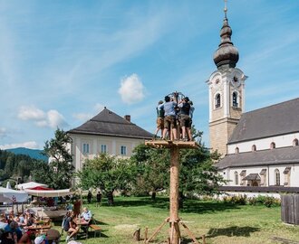 Männer in Tracht stehen auf einem Kletterbaum beim Fest im Pfarrhof Altenmarkt, Zuschauer sitzen auf Bierbänken im Grünen. | © Matthias Fritzenwallner