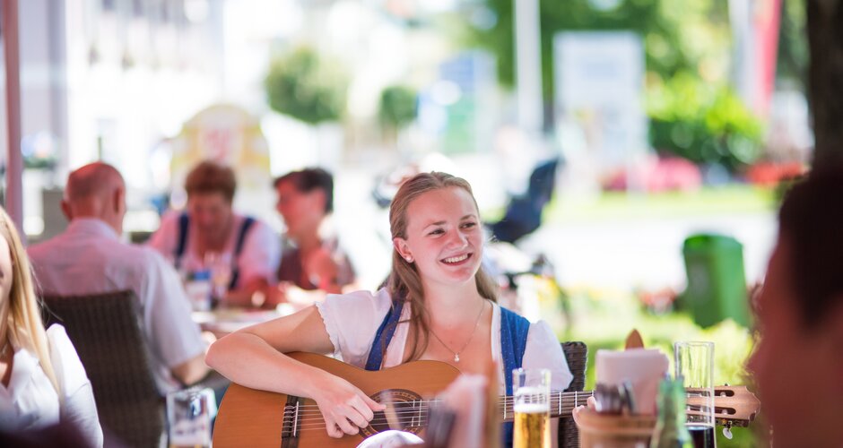 Smiling young woman in dirndl playing guitar at Musikanten.Roas, people sit together at tables with drinks | © Nadia Jabli Photography