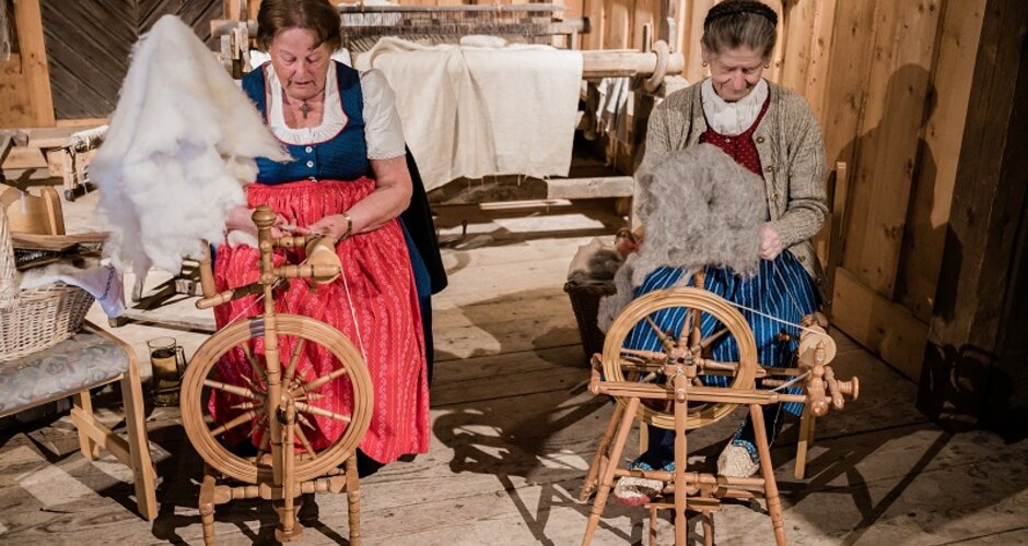 Two women in traditional dress spin sheep’s wool at spinning wheels during a heritage craft event in a rustic wooden room. | © Matthias Fritzenwallner