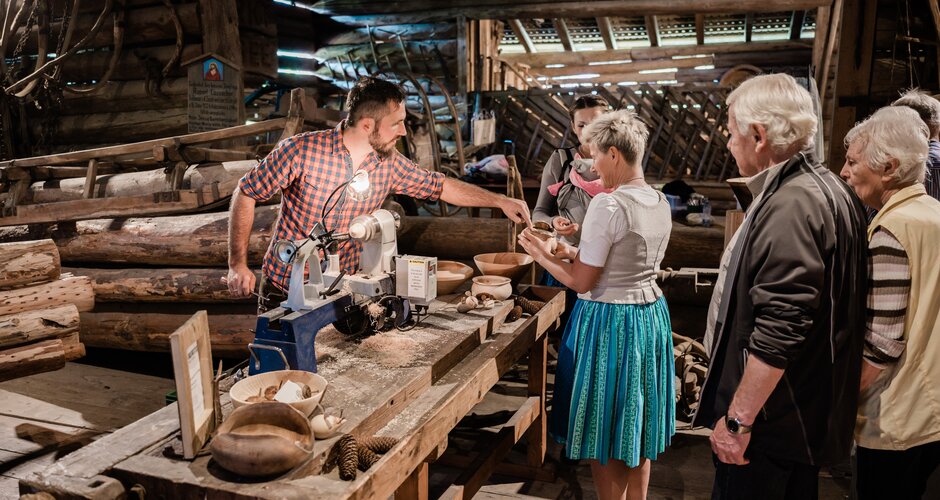 Craftsman shows woodturning to visitors, group watches traditional handicraft in rustic workshop during heritage event. | © Matthias Fritzenwallner