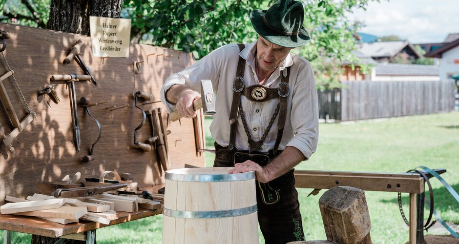 Barrel maker in leather trousers works on wooden cask using hammer, surrounded by traditional tools at craft demonstration. | © Matthias Fritzenwallner
