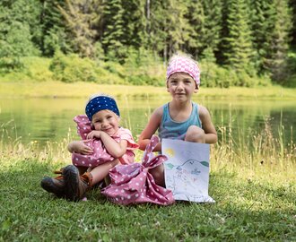 Zwei Kinder sitzen am See beim Event See.Musik, mit Picknickbeuteln und einem selbstgemalten Bild mit Sonne und Bergen. | © Matthias Fritzenwallner