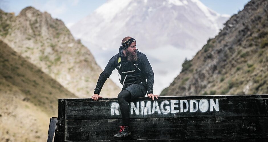 Ein bärtiger Teilnehmer in schwarzer Sportkleidung überklettert ein Hindernis beim Runmageddon vor alpiner Bergkulisse. | © Runmageddon X_Filzmoos_Austria_©AdamRyszczyk_www.runmageddon-austria.at