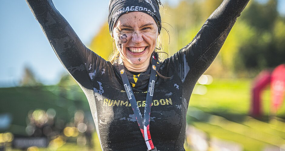 Strahlende Finisherin mit Stirnband, Schlamm im Gesicht und Finisher-Medaille jubelt im Zielbereich des Runmageddon-Events. | © Runmageddon X_Filzmoos_Austria_©AdamRyszczyk_www.runmageddon-austria.at