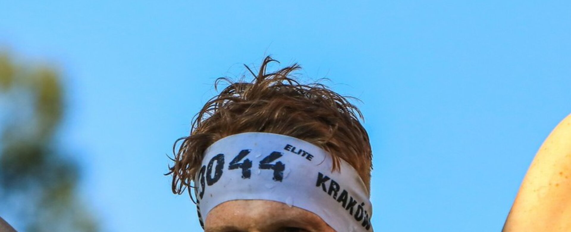 Ein erschöpfter, aber strahlender Teilnehmer hält am Ziel des Runmageddon ein gelbes Zielflaggenband über dem Kopf hoch. | © Adam Ryszczyk