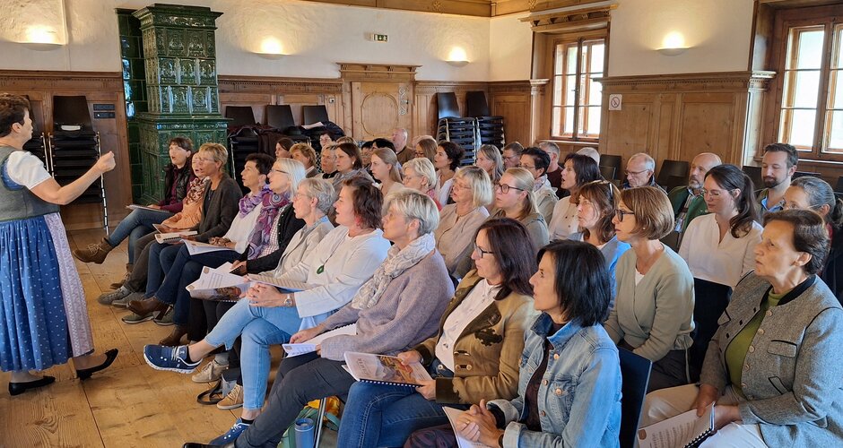 Conductor leads choir rehearsal with participants in wood-paneled hall | © Flachau Tourismus