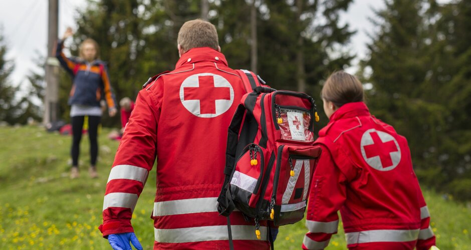 Zwei Sanitäter vom Österreichischen Roten kreuz gehen zu einer winkenden Person vor einem Gipfelkreuz | © OERK - Thomas Holly Kellner