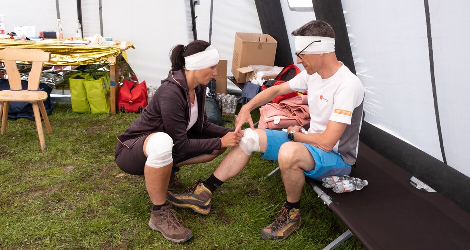 Eine Frau mit Verband am Knie und am Kopf verbindet einem Mann mit Verband am Kopf das Knie