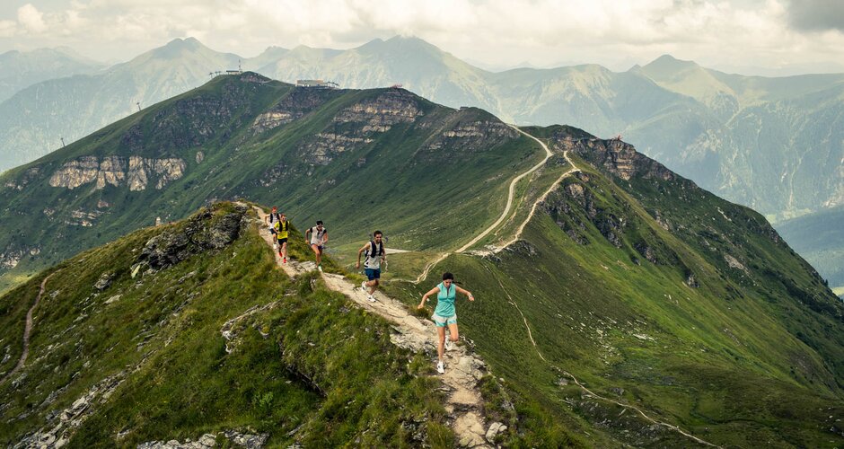 Several trail runners running along a narrow alpine ridge with green mountain peaks and a cloudy sky in the background | © Gasteinertal Tourismus GmbH, Michael Mueller