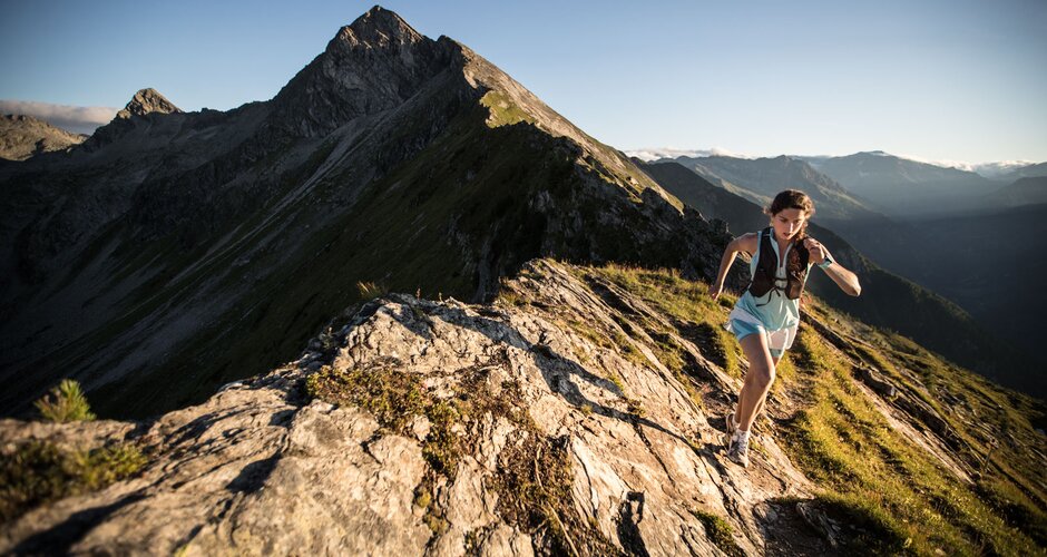 Female trail runner on a rocky ridge path with sunlight and impressive mountain scenery in the background | © Gasteinertal Tourismus GmbH, Michael Mueller