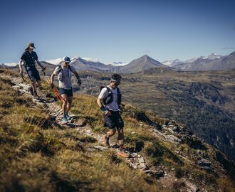 Three trail runners hiking along a rocky mountain path with green valleys and snowy High Tauern peaks in the background | © Stefan Schaaf PEAK ART IMAGES